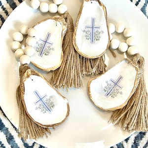 May include: Four oyster shell ornaments with gold trim and a cross design. Each ornament has a brown tassel and is strung on a white bead garland.