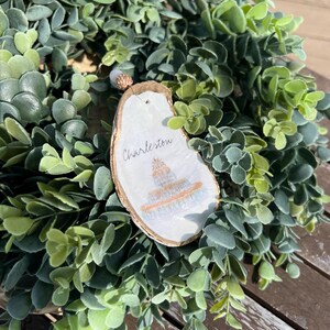 Op de afbeelding: Een witte oester schelp ornament met gouden verf en een handgeschilderd beeld van een gebouw. De ornament is versierd met een witte parel en de tekst "Charleston" is op de schelp geschreven.