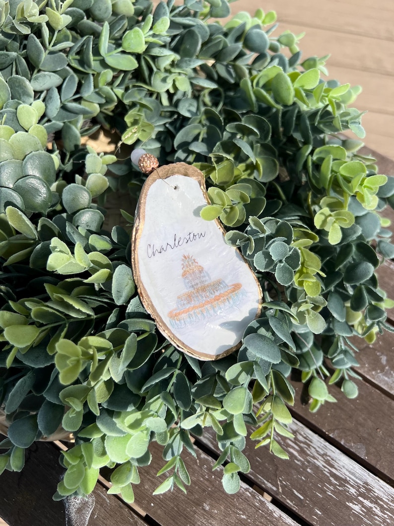Op de afbeelding: Een witte oester schelp ornament met gouden verf en een handgeschilderd beeld van een gebouw. De ornament is versierd met een witte parel en de tekst "Charleston" is op de schelp geschreven.