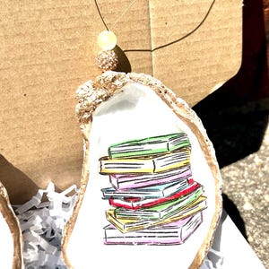 May include: A white oyster shell with a gold rim, painted with a stack of colorful books. The oyster shell is hanging from a gold string with a sparkly bead.