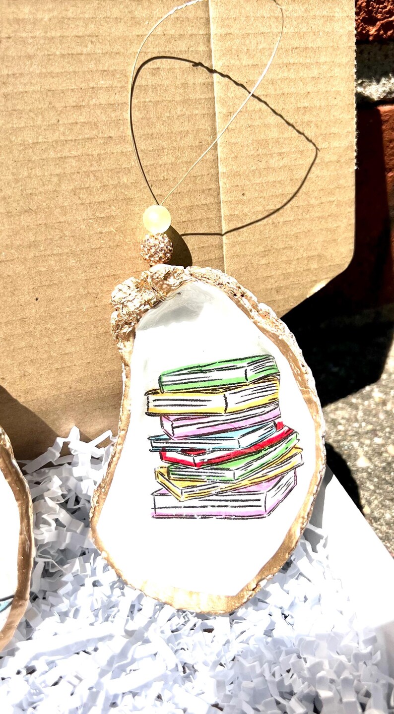 May include: A white oyster shell with a gold rim, painted with a stack of colorful books. The oyster shell is hanging from a gold string with a sparkly bead.
