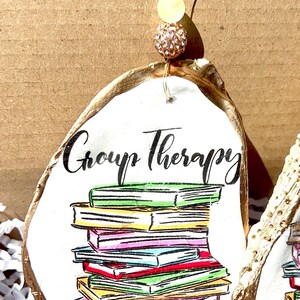 May include: A white oyster shell ornament with gold trim, featuring a stack of colorful books and the text "Group Therapy".