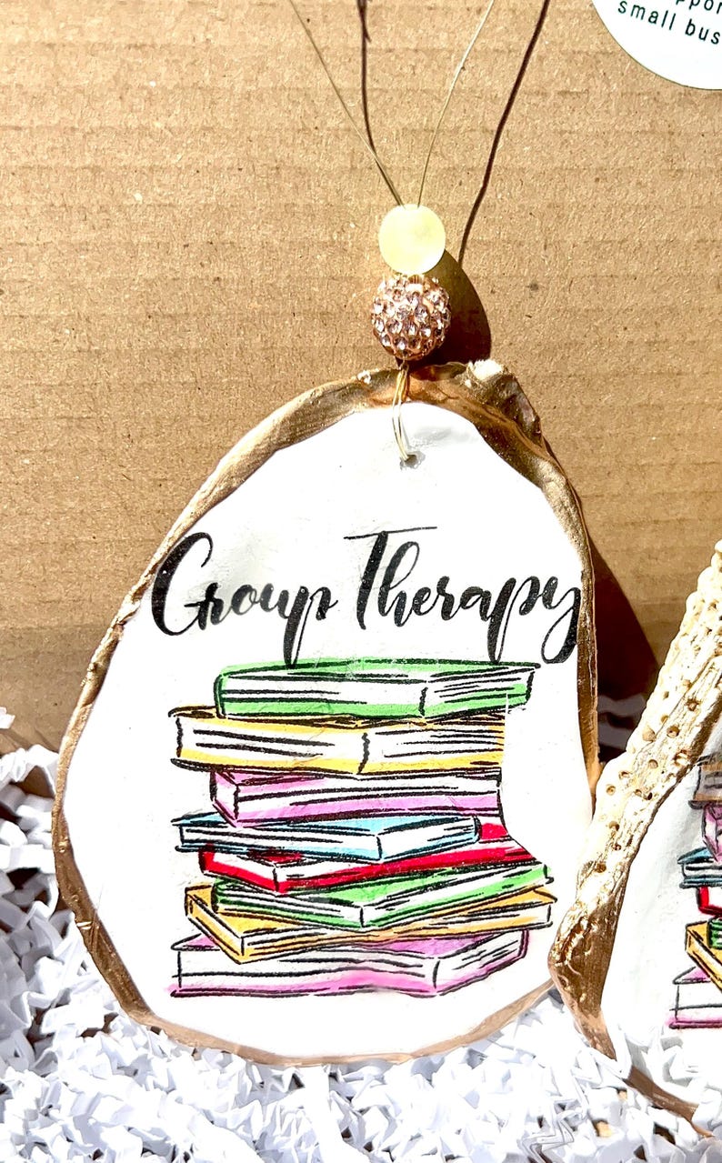 May include: A white oyster shell ornament with gold trim, featuring a stack of colorful books and the text "Group Therapy".