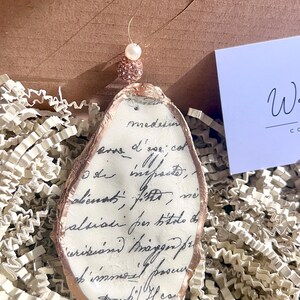 以下が含まれることがあります： 黒インクの手書き文字が入ったカキ殻のオーナメント。 殻は金色の縁取りがあり、パールとビーズのアクセントが付いた金のワイヤーで吊り下げられています。 オーナメントは白いクリンクル紙の上に飾られています。