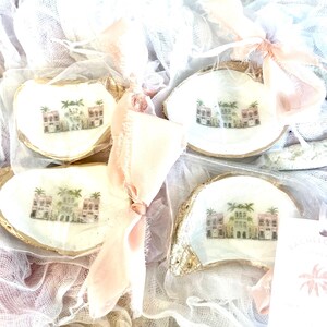 May include: Four white oyster shell trinket dishes with gold trim. Each dish features a painted scene of a row of colorful houses with palm trees. The dishes are displayed on a white and pink fabric background.