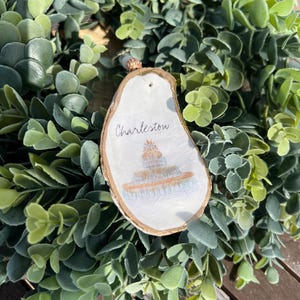 Op de afbeelding: Een wit houten ornament met een gouden rand, met een handgeschilderde illustratie van een gebouw met het woord "Charleston" erboven.