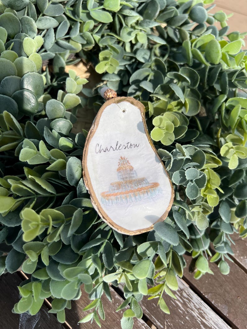 Op de afbeelding: Een wit houten ornament met een gouden rand, met een handgeschilderde illustratie van een gebouw met het woord "Charleston" erboven.