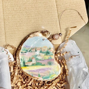 Op de afbeelding: Een handgeschilderd oester schelp ornament met een landschapssc&egrave;ne met een dorp, bomen en lavendelvelden. Het ornament is verguld en heeft een gouden koord om op te hangen.