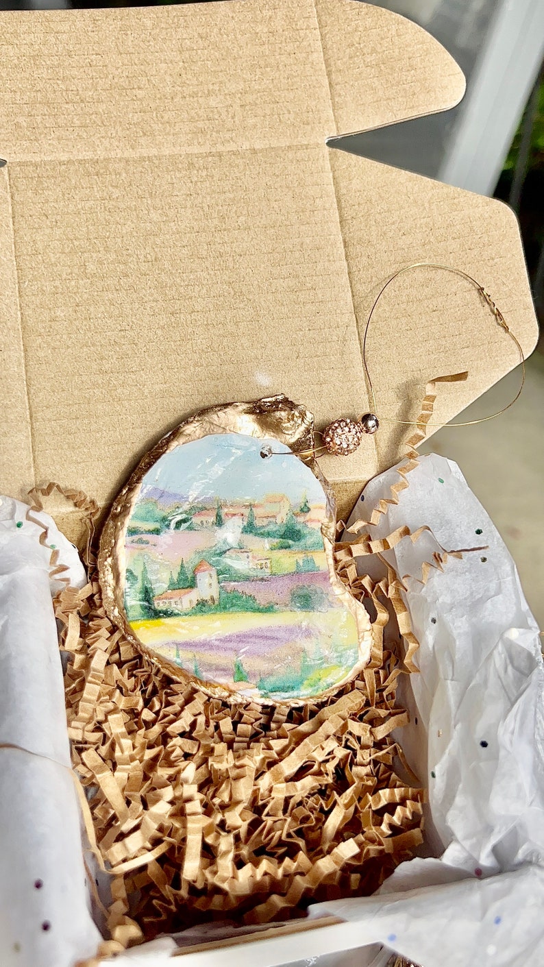 Op de afbeelding: Een handgeschilderd oester schelp ornament met een landschapssc&egrave;ne met een dorp, bomen en lavendelvelden. Het ornament is verguld en heeft een gouden koord om op te hangen.