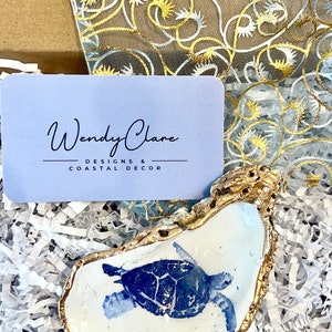 May include: A white ceramic oyster shell dish with a gold rim, featuring a blue and white painted sea turtle design. The dish is sitting on white shredded paper.