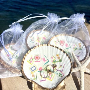 May include: A seashell ring holder with gold trim, holding two gold wedding rings. The shell is decorated with colorful Mahjong tile designs. Several similar ring holders are in sheer white bags, with a starfish nearby.
