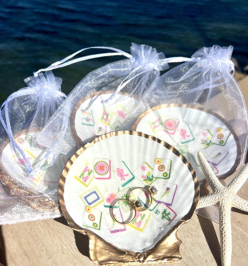 May include: A seashell ring holder with gold trim, holding two gold wedding rings. The shell is decorated with colorful Mahjong tile designs. Several similar ring holders are in sheer white bags, with a starfish nearby.