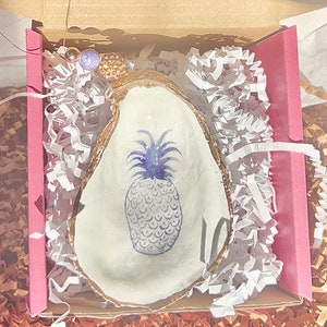 May include: A white ceramic oyster shell dish with a gold rim, featuring a blue and white pineapple design. The dish is nestled in white and brown shredded paper inside a pink and brown box.