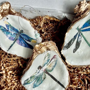 May include: Three gold-rimmed oyster shell ornaments with dragonfly designs. Each shell features a different dragonfly with intricate details and vibrant colors, including blue, green, and red.
