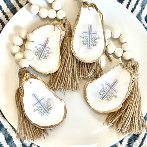 May include: Three white oyster shells with gold trim and a blue cross design with floral accents. Each shell has a brown tassel attached and is strung on a white wooden bead garland.