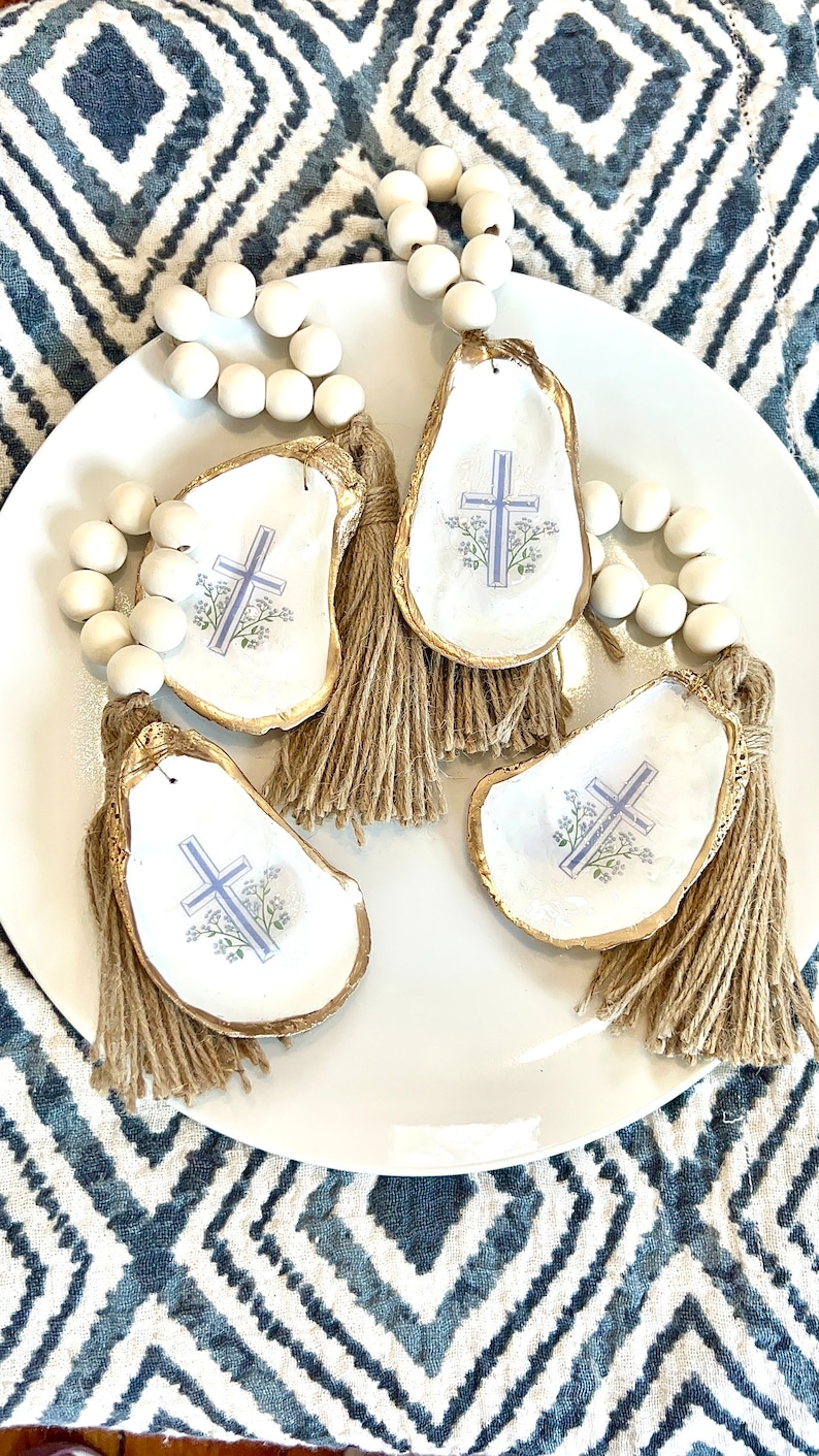 May include: Three white oyster shells with gold trim and a blue cross design with floral accents. Each shell has a brown tassel attached and is strung on a white wooden bead garland.