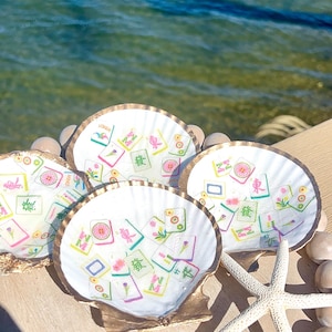 May include: Four white scallop shells with gold rims, each decorated with colorful Mahjong tile designs. A starfish and small stones are next to the shells. The background shows the ocean.
