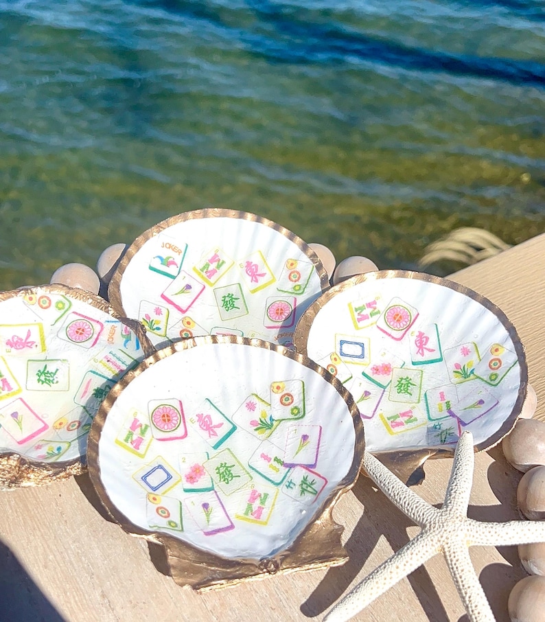 May include: Four white scallop shells with gold rims, each decorated with colorful Mahjong tile designs. A starfish and small stones are next to the shells. The background shows the ocean.