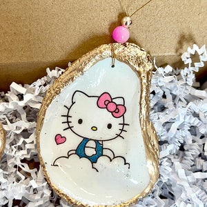 Może przedstawiać: Biała ozdoba w kształcie muszli ostrygi z pozłacanym brzegiem, przedstawiająca kreskówkową Hello Kitty w niebieskim stroju i różową kokardką. Ozdoba jest zawieszona na złotej sznurce z różową koralikiem.