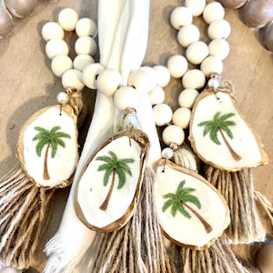 May include: Set of four oyster shell napkin rings with gold trim and a palm tree design. Each ring has a jute tassel. The rings are strung on a white wooden bead garland.