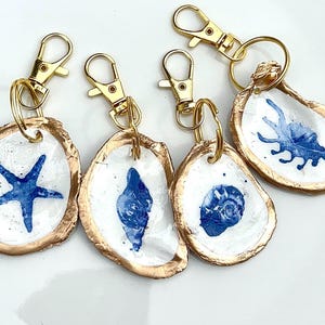 May include: Four oyster shell keychains featuring hand-painted blue marine life designs including a starfish, a conch shell, a spiral shell, and a crab. Each shell has a gold-painted edge and is attached to a gold-tone metal lobster clasp.