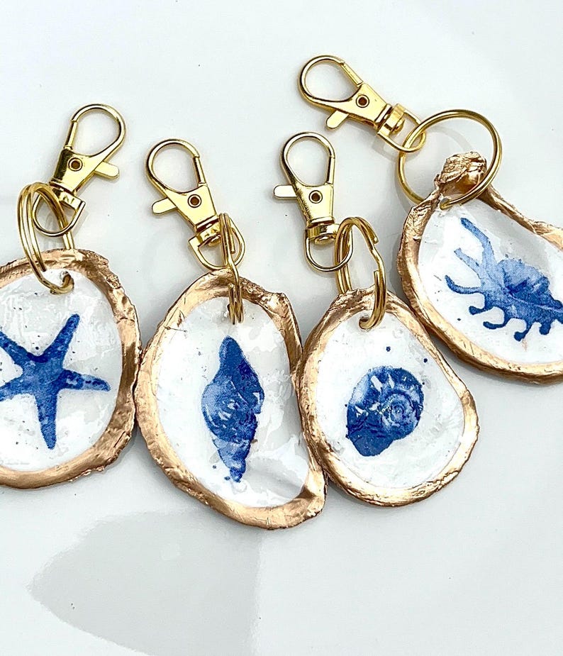 May include: A collection of five keychains featuring painted oyster shells. Each shell is painted with a different blue sea creature design, including a starfish and various shells. The shells have gold-colored edges and are attached to gold-colored keychains.