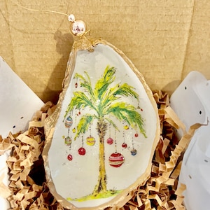 May include: A gold-painted oyster shell ornament with a painted palm tree and colorful ornaments hanging from the branches. The ornament is nestled in brown packing paper.