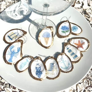 May include: Oyster shell wine charms with gold edging and beach-themed watercolour designs. The charms depict an ice cream cone, lobster, and a blue swimsuit. A wine glass is positioned above the charms, all on a white plate.