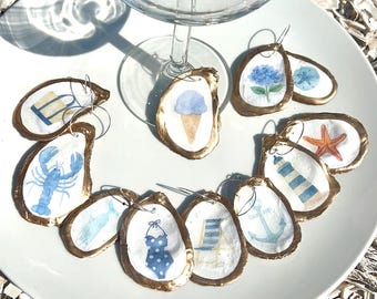 Könnte beinhalten: Weinglas-Anhänger aus Austernschalen mit goldenen Rändern und Strand-Aquarelldesigns. Die Anhänger zeigen Eiswaffel, Hummer und einen blauen Badeanzug. Ein Weinglas befindet sich über den Anhängern, auf einem weißen Teller.