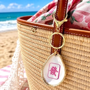 May include: A gold-colored keychain with a white, teardrop-shaped charm featuring a pink Chinese character. The charm hangs from a gold ring and clasp attached to a woven straw tote bag with brown leather trim. A floral scarf is visible inside the bag.