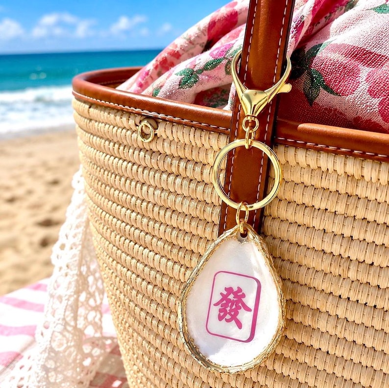 May include: A gold-colored keychain with a white, teardrop-shaped charm featuring a pink Chinese character. The charm hangs from a gold ring and clasp attached to a woven straw tote bag with brown leather trim. A floral scarf is visible inside the bag.