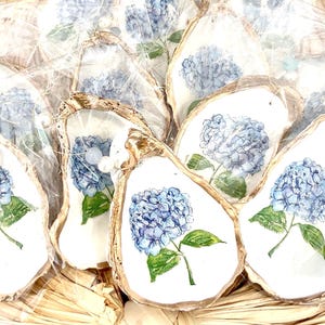 K&ouml;nnte beinhalten: Eine Gruppe von Austernmuschel-Anh&auml;ngern, auf denen jeweils eine blaue Hortensienbl&uuml;te gemalt ist. Die Muscheln sind in einem Korb angeordnet und in durchsichtige Plastikfolie verpackt.