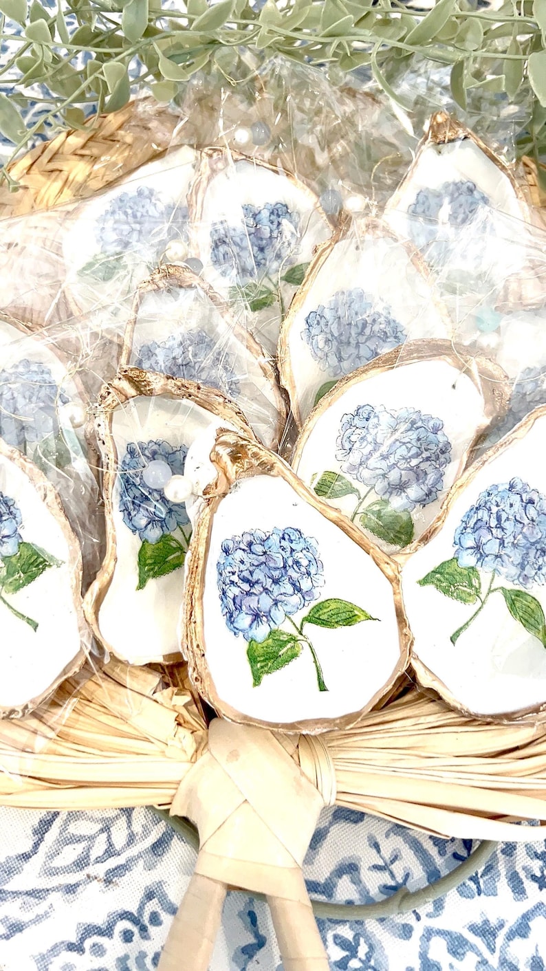 K&ouml;nnte beinhalten: Eine Gruppe von Austernmuschel-Anh&auml;ngern, auf denen jeweils eine blaue Hortensienbl&uuml;te gemalt ist. Die Muscheln sind in einem Korb angeordnet und in durchsichtige Plastikfolie verpackt.