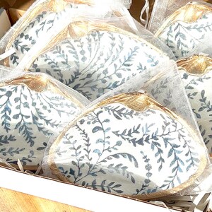 May include: A box filled with several decorative clam shells. Each shell has a white background with a blue floral pattern and a gold-colored trim. The shells are individually wrapped in sheer white bags.