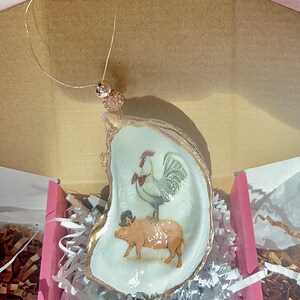 May include: A gold-painted oyster shell ornament with a rooster and pig illustration. The rooster is wearing a red bow tie and the pig is wearing a black top hat.