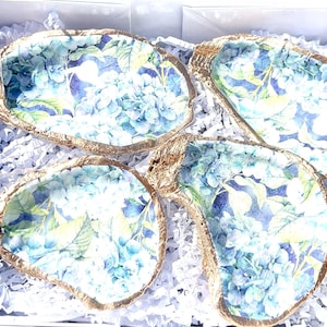May include: Four decorative oyster shells with a blue and green floral pattern and gold trim. The shells are arranged in a white shredded paper box.