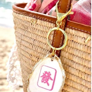 May include: A gold-colored keychain with a white and pink Mahjong tile charm is attached to a brown leather strap on a woven straw tote bag. The charm has a gold-colored edge and a pink Chinese character.