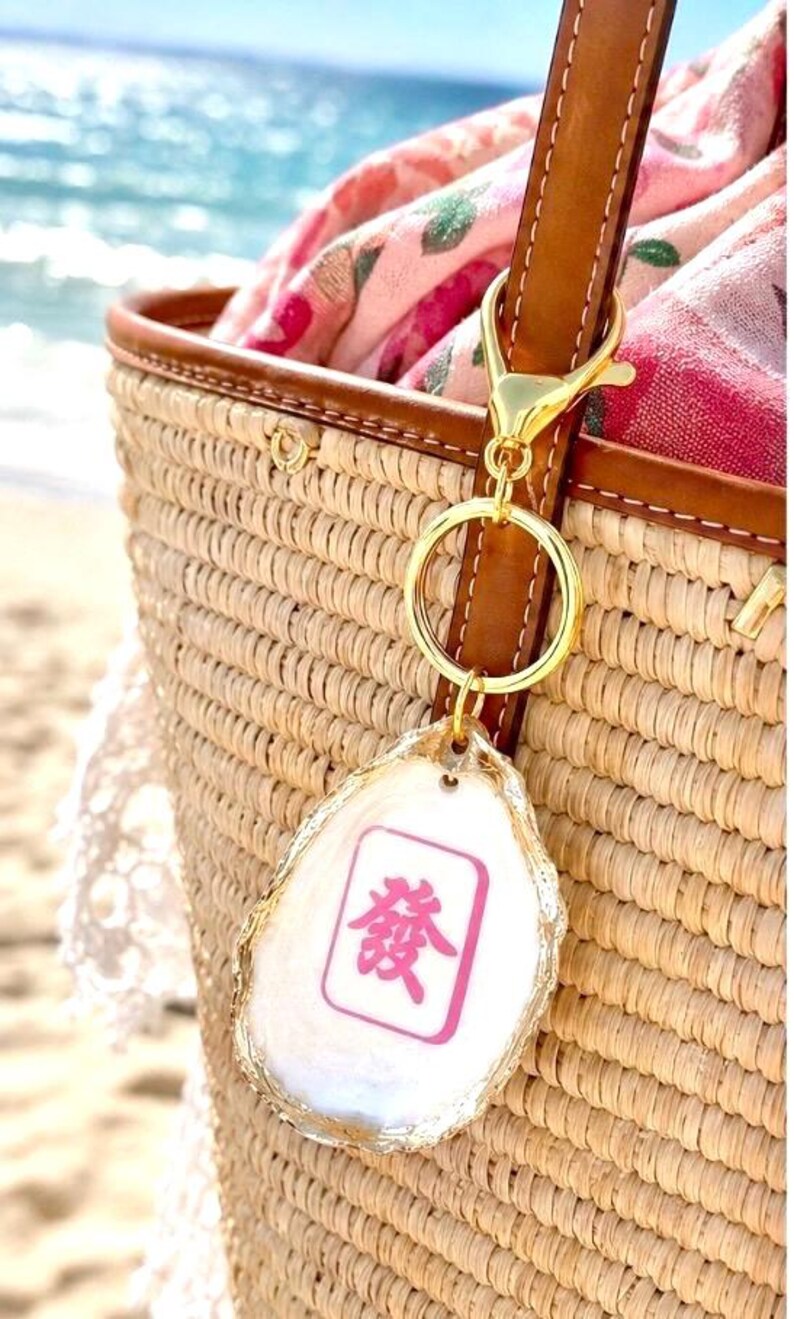 May include: A gold-colored keychain with a white and pink Mahjong tile charm is attached to a brown leather strap on a woven straw tote bag. The charm has a gold-colored edge and a pink Chinese character.