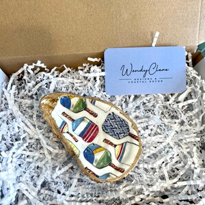 May include: A white oyster shell with a gold rim, painted with colorful paddleball rackets and a blue and white geometric pattern. The shell is sitting in white shredded paper inside a brown cardboard box. The box also contains a blue card with the text "Wendy Clare Designs & Coastal Decor".
