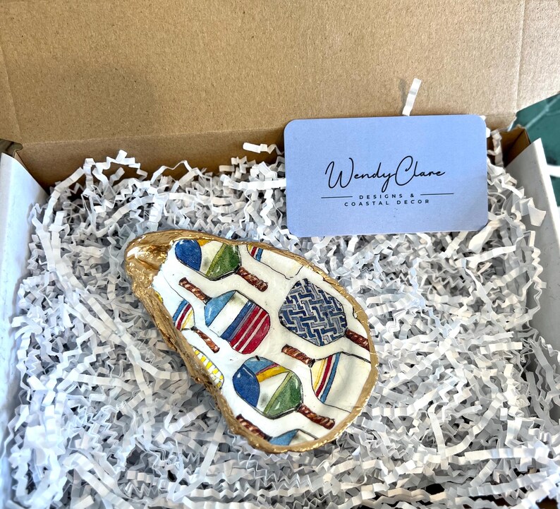 May include: A white oyster shell with a gold rim, painted with colorful paddleball rackets and a blue and white geometric pattern. The shell is sitting in white shredded paper inside a brown cardboard box. The box also contains a blue card with the text "Wendy Clare Designs & Coastal Decor".
