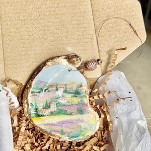 Op de afbeelding: Een handgeschilderd oester schelp ornament met een landschapssc&egrave;ne met een dorp, bomen en lavendelvelden. Het ornament is verguld en heeft een klein, helder kristal versiering. Het is verpakt in een bruine doos met gescheurd papier.