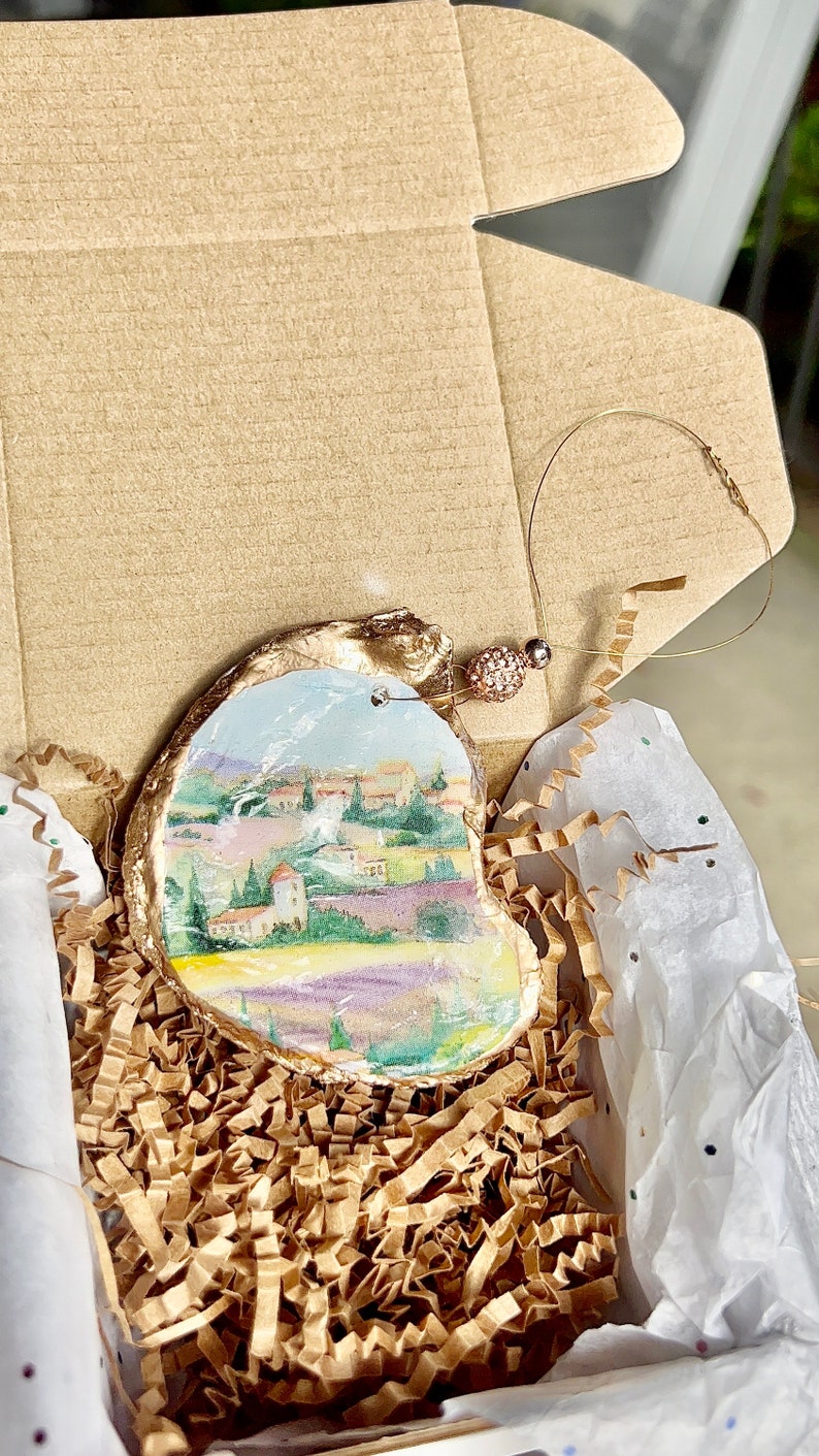 Op de afbeelding: Een handgeschilderd oester schelp ornament met een landschapssc&egrave;ne met een dorp, bomen en lavendelvelden. Het ornament is verguld en heeft een klein, helder kristal versiering. Het is verpakt in een bruine doos met gescheurd papier.