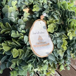 Op de afbeelding: Een wit houten ornament met een geschilderd beeld van een gebouw en de tekst "Charleston". Het ornament hangt aan een gouden touwtje met een witte kraal.