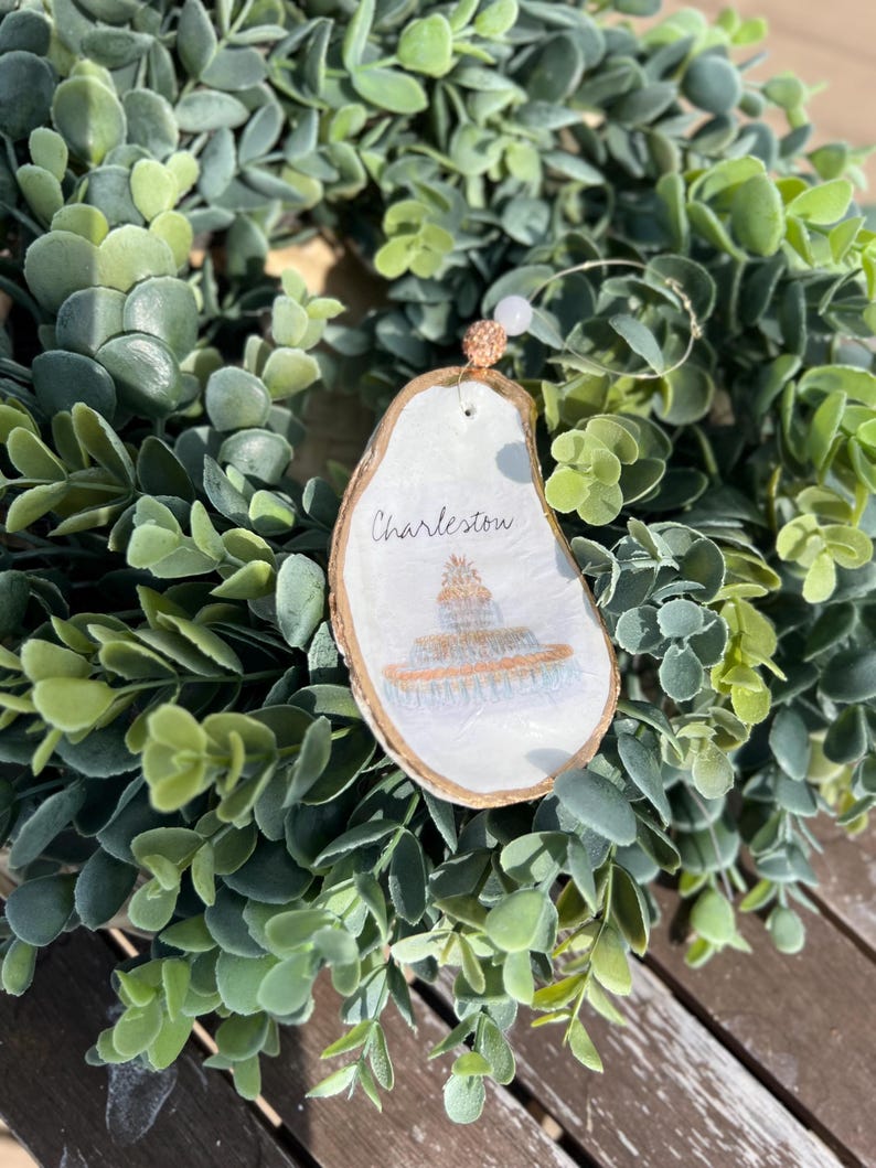 Op de afbeelding: Een wit houten ornament met een geschilderd beeld van een gebouw en de tekst "Charleston". Het ornament hangt aan een gouden touwtje met een witte kraal.