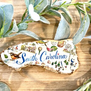 Op de afbeelding: Een wit en goud geschilderde houten omtrek van de staat South Carolina met verschillende illustraties van bezienswaardigheden en activiteiten, waaronder een watermeloen, een golfbal, een reuzenrad, een vuurtoren en een parasol. De tekst "South Carolina" is geschreven in blauwe cursieve letters.