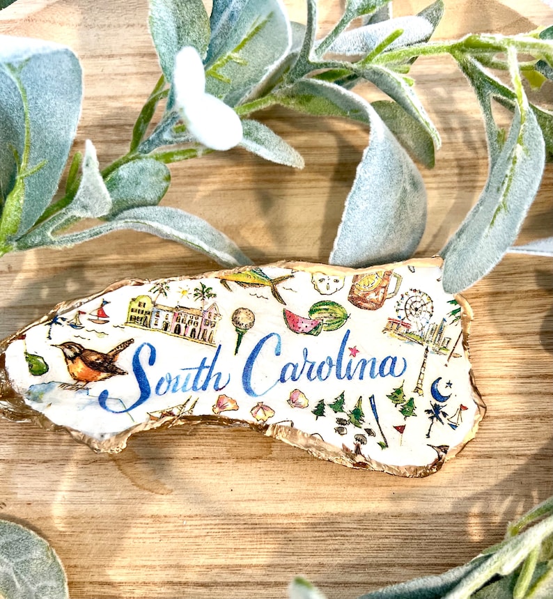 Op de afbeelding: Een wit en goud geschilderde houten omtrek van de staat South Carolina met verschillende illustraties van bezienswaardigheden en activiteiten, waaronder een watermeloen, een golfbal, een reuzenrad, een vuurtoren en een parasol. De tekst "South Carolina" is geschreven in blauwe cursieve letters.