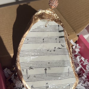 May include: Gold-painted oyster shell ornament with a sheet of music printed on the inside. The ornament is hanging from a thin gold string with a small pink and gold bead.