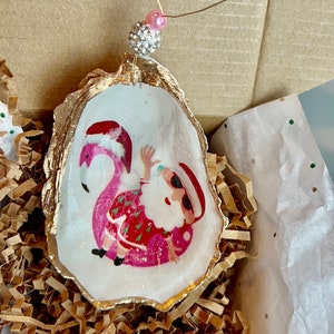 May include: A gold-painted oyster shell ornament with a white background featuring a cartoon image of Santa Claus riding a pink flamingo. The ornament has a pink and white beaded string for hanging.