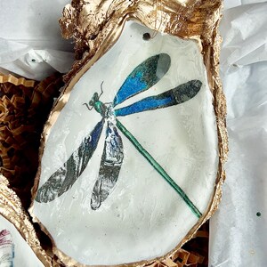 May include: A gold-painted oyster shell with a blue and green dragonfly design. The shell is sitting on a bed of brown packing paper.