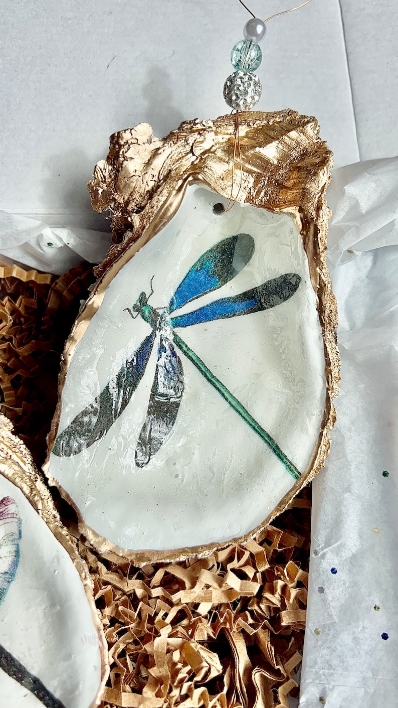 May include: A gold-painted oyster shell with a blue and green dragonfly design. The shell is sitting on a bed of brown packing paper.